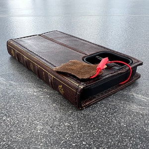 May include: A brown leather phone case designed to look like a vintage book. The case has gold-colored text and accents, with a flap and a red string attached. The case is designed to hold a phone.