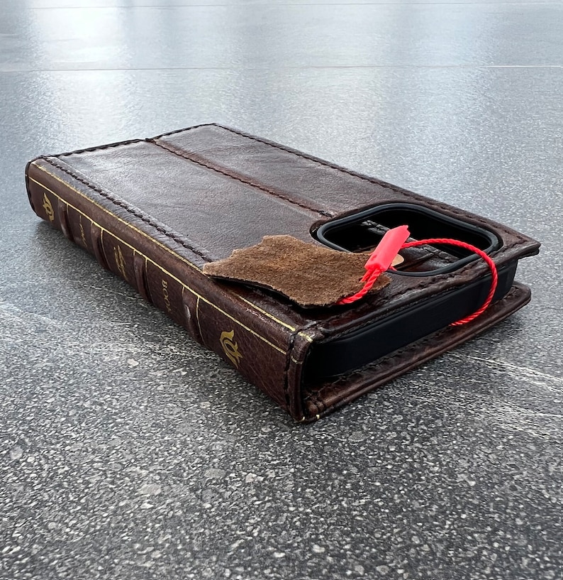 May include: A brown leather phone case designed to look like a vintage book. The case has gold-colored text and accents, with a flap and a red string attached. The case is designed to hold a phone.