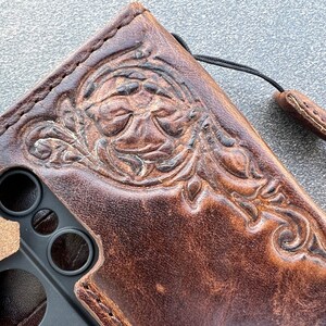 May include: Brown leather phone case with an embossed floral design. The case has a distressed finish and a black lanyard attached. The design features detailed leaves and swirls, adding a touch of elegance to the protective accessory.