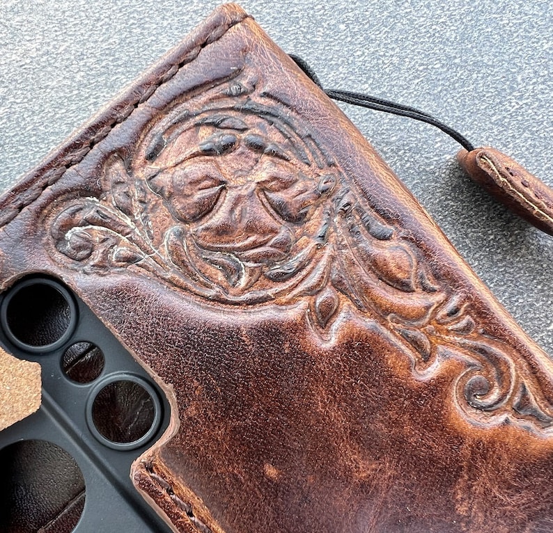 May include: Brown leather phone case with an embossed floral design. The case has a distressed finish and a black lanyard attached. The design features detailed leaves and swirls, adding a touch of elegance to the protective accessory.