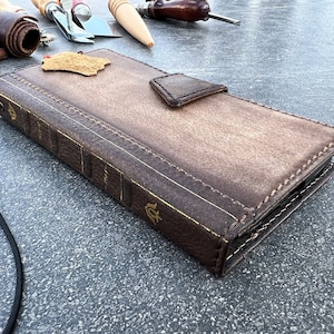 Könnte beinhalten: Eine braune Leder-Handyhülle mit einem buchähnlichen Design. Die Hülle hat goldene Akzente und Nähte. Die Hülle liegt auf einer grauen Oberfläche.
