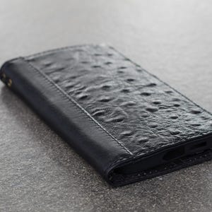 May include: A black leather phone case with a textured, ostrich-like pattern. The case has a stitched border and a wrist strap attached. The case is designed to hold a phone and protect it from damage.