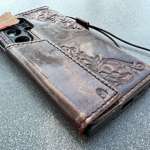 May include: A brown leather phone case with an embossed floral design. The case has a distressed finish and a magnetic closure. A black lanyard is attached to the case.
