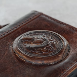 Könnte beinhalten: Eine braune Ledergeldbörse mit einem geprägten Pferdekopf-Emblem. Die Geldbörse hat ein abgenutztes, strukturiertes Aussehen mit sichtbaren Nähten und einem sicheren Verschluss. Das Design deutet auf einen klassischen, rustikalen Stil hin.