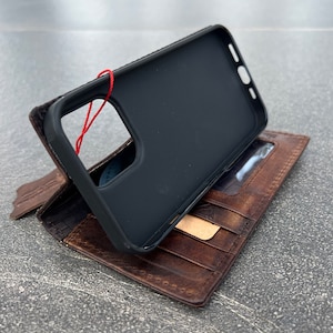 May include: A black phone case with a red string attached, resting on top of a brown leather wallet. The wallet has multiple card slots and a clear ID window. The leather has a distressed appearance.
