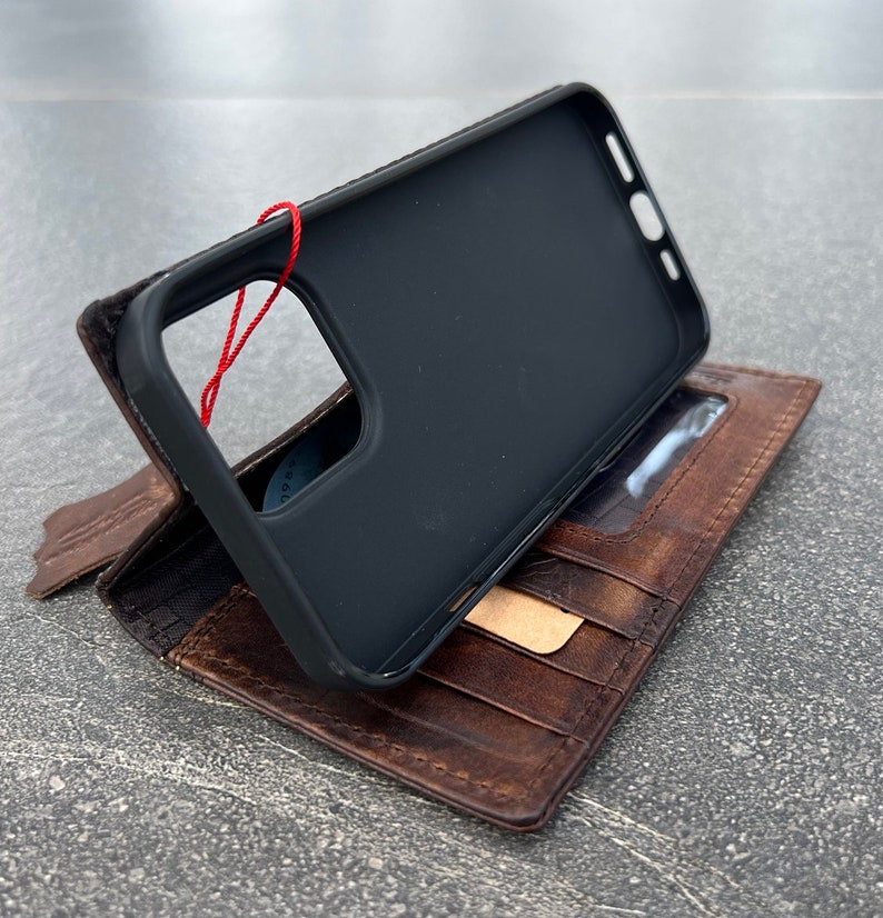 May include: A black phone case with a red string attached, resting on top of a brown leather wallet. The wallet has multiple card slots and a clear ID window. The leather has a distressed appearance.