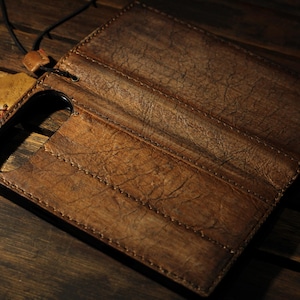 May include: A brown leather phone case with a textured surface and stitched details. The case is open, revealing the interior and a cutout for the phone's camera. A leather tag and black cord are attached.