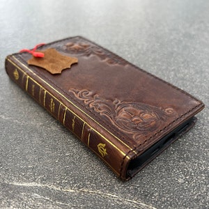 May include: A brown leather phone case with a book-like design. The case features embossed floral patterns and gold-colored accents on the spine, with the word "BOOK" visible. A small leather tag is attached.