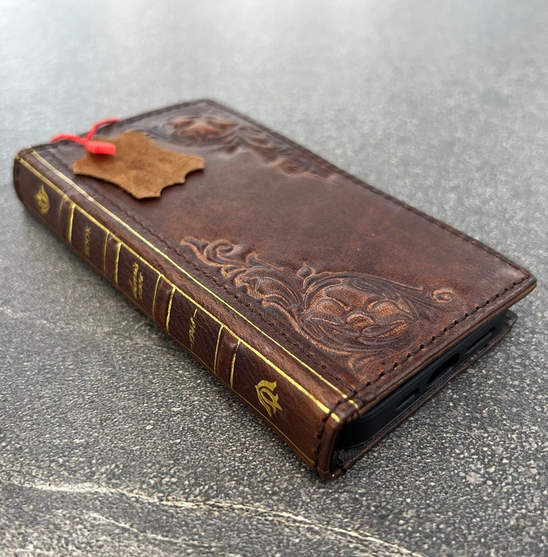 May include: A brown leather phone case with a book-like design. The case features embossed floral patterns and gold-colored accents on the spine, with the word "BOOK" visible. A small leather tag is attached.