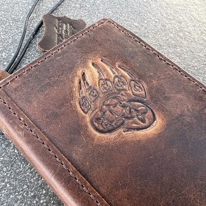 May include: A brown leather wallet with a bear paw design embossed on the front. The wallet has a stitched edge and a distressed look.