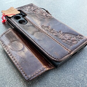 May include: A brown leather phone case with an embossed floral design. The case is open, revealing the phone's camera and screen. A small leather tag is attached with a red string. The case has a wallet-style design.