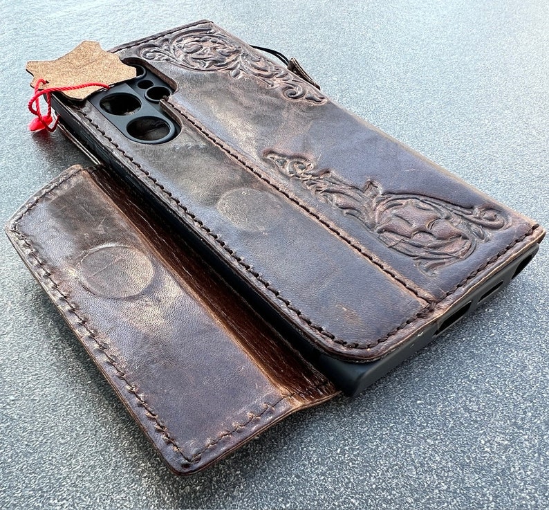 May include: A brown leather phone case with an embossed floral design. The case is open, revealing the phone's camera and screen. A small leather tag is attached with a red string. The case has a wallet-style design.