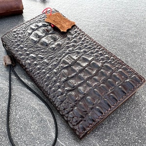 May include: A brown leather wallet with a crocodile skin pattern and a black strap. The wallet is closed and has a small piece of leather attached to it.