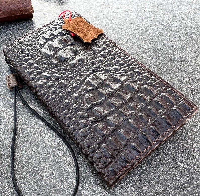 May include: A brown leather wallet with a crocodile skin pattern and a black strap. The wallet is closed and has a small piece of leather attached to it.