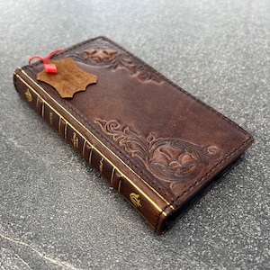 May include: A brown leather wallet with embossed floral designs and gold-colored edges. The wallet has a book-like appearance with text on the spine and a small leather tag attached. The overall design is vintage and detailed.