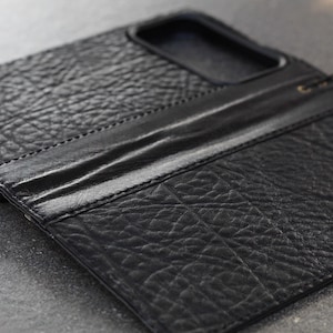 Könnte beinhalten: Schwarze Leder-Handyhülle mit strukturierter Oberfläche. Die Hülle hat ein Flip-Cover-Design und einen Ausschnitt für die Kamera. Die Nähte sind an den Kanten sichtbar, und ein kleiner Riemen ist befestigt.