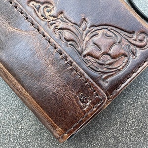 May include: Close-up of a brown leather wallet with a floral embossed design. The wallet has a distressed appearance with visible stitching and a black cord attached. The leather shows signs of wear, adding to its rustic charm.