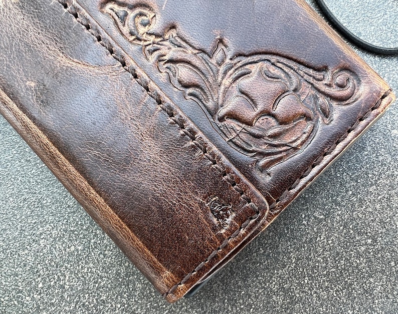 May include: Close-up of a brown leather wallet with a floral embossed design. The wallet has a distressed appearance with visible stitching and a black cord attached. The leather shows signs of wear, adding to its rustic charm.