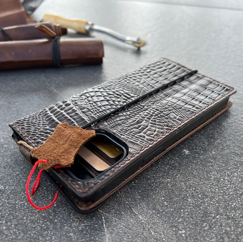 May include: A brown leather phone case with a crocodile pattern. The case has a flap that closes with a strap and a small leather tag attached to it. There are also slots for credit cards inside the case.