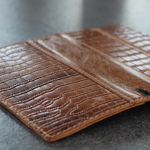 Può includere: Una custodia per telefono in pelle marrone con una texture a effetto pelle di coccodrillo. La custodia è aperta, rivelando l'interno e un telefono nero. La pelle ha una tonalità calda e cuciture visibili.