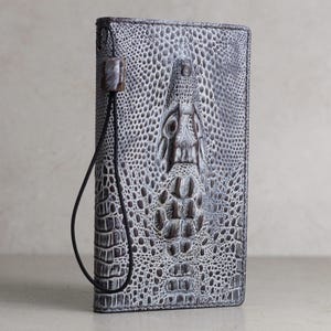 Könnte beinhalten: Eine graue Brieftasche aus Alligatorleder mit einem schwarzen Riemen. Die Brieftasche hat ein Krokodilkopf-Design auf der Vorderseite.