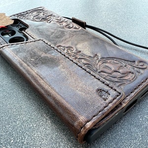 May include: A brown leather phone case with an embossed floral design. The case has a wallet-style flap and a black lanyard. The leather has a distressed finish, and a small tag is attached.