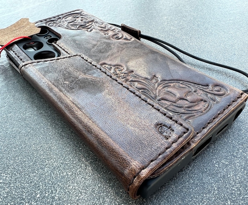 May include: A brown leather phone case with an embossed floral design. The case has a wallet-style flap and a black lanyard. The leather has a distressed finish, and a small tag is attached.