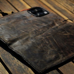 Puede incluir: Una funda de teléfono de cuero marrón desgastado con bordes cosidos. La funda tiene una abertura para la cámara y un pequeño lazo. El cuero tiene un aspecto desgastado y texturizado, lo que sugiere un estilo vintage. La funda está abierta, revelando el interior.