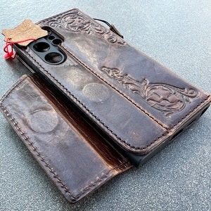 May include: A brown leather phone case with an embossed floral design. The case is open, revealing the phone's camera and screen area. A small leather tag is attached with a red string.