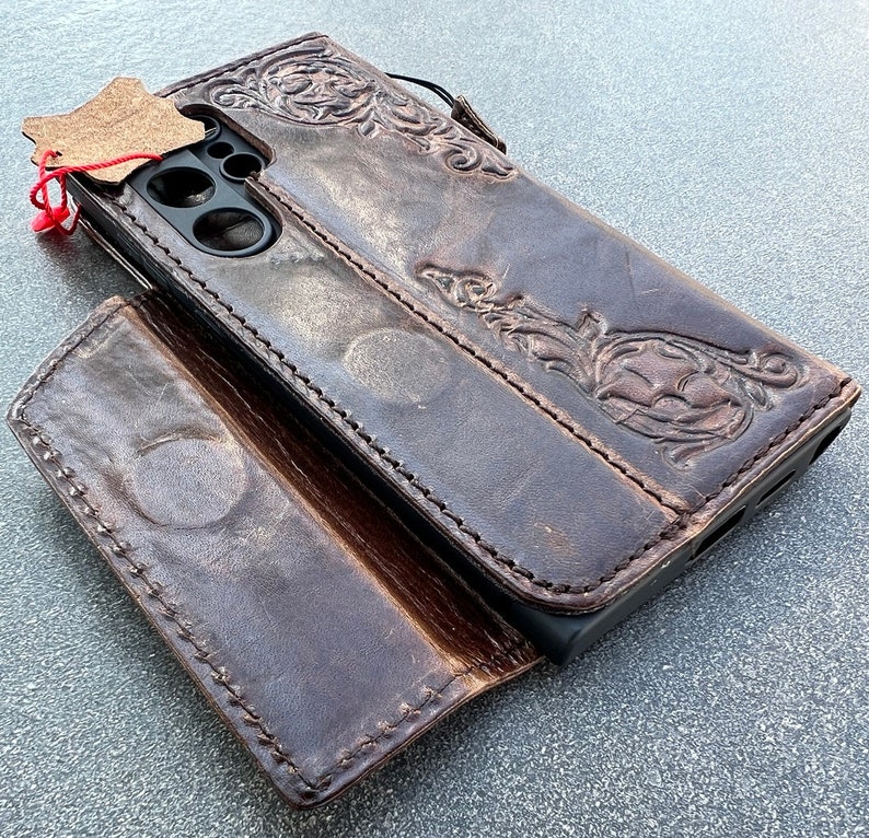 May include: A brown leather phone case with an embossed floral design. The case is open, revealing the phone's camera and screen area. A small leather tag is attached with a red string.
