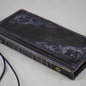 May include: A black leather wallet with a gold embossed design and a black strap. The wallet has a vintage look and is closed.