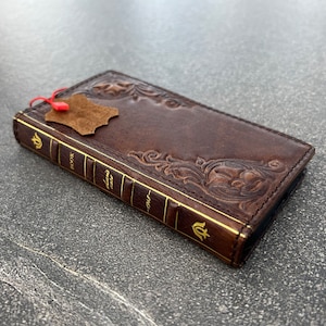May include: A brown leather phone case with embossed floral designs and gold accents. The case is shaped like a book, with the word "BOOK" and "1948" printed on the spine. A small leather tag hangs from the top.