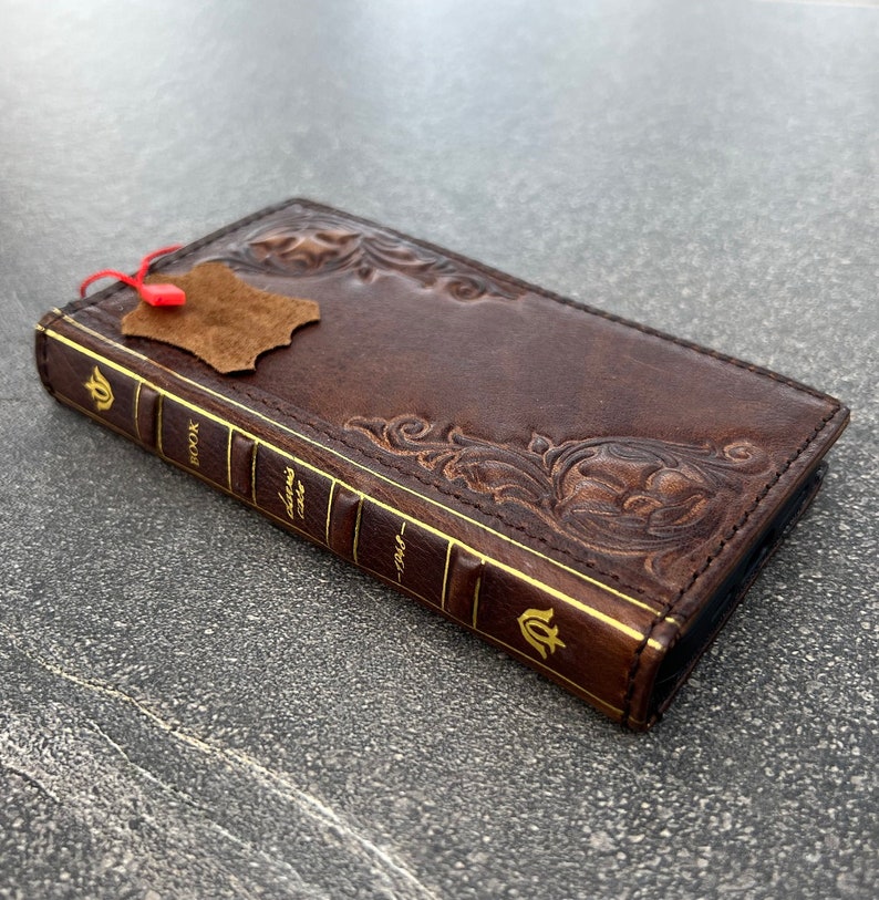 May include: A brown leather phone case with embossed floral designs and gold accents. The case is shaped like a book, with the word "BOOK" and "1948" printed on the spine. A small leather tag hangs from the top.