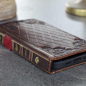 May include: A brown leather phone case designed to look like a vintage book. The case features embossed floral and diamond patterns, with gold-colored accents along the spine and a red tab closure. The case is designed to fit a smartphone.