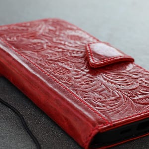 May include: A red leather phone case with an embossed floral pattern. The case features a magnetic clasp and a wrist strap. The stitching is visible, and the case appears to be made of high-quality materials.
