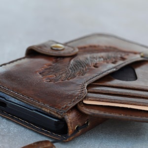Peut inclure: Portefeuille en cuir marron avec un motif d'aigle en relief. Le portefeuille est ouvert, révélant des fentes pour cartes et un étui de téléphone noir. Le cuir a un aspect vieilli et se ferme avec un bouton-pression.