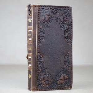 May include: A dark brown leather book cover with embossed floral designs. The spine features gold lettering and a decorative emblem. The cover has a clasp closure and is designed to protect a book. The overall aesthetic is vintage and elegant.