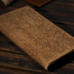 Puede incluir: Una cartera rectangular de cuero marrón claro con una superficie texturizada. La cartera está cerrada. El fondo es una superficie de madera oscura.