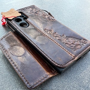 May include: Dark brown leather phone case with embossed floral and scrollwork design. The case is open, revealing the phone's camera and screen. A small leather tag is attached with a red string.
