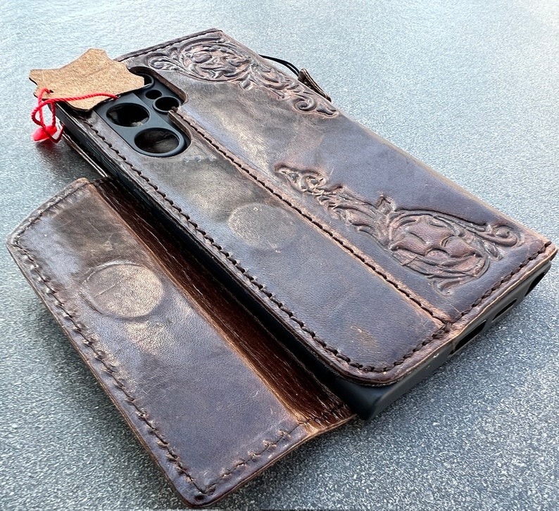 May include: Dark brown leather phone case with embossed floral and scrollwork design. The case is open, revealing the phone's camera and screen. A small leather tag is attached with a red string.