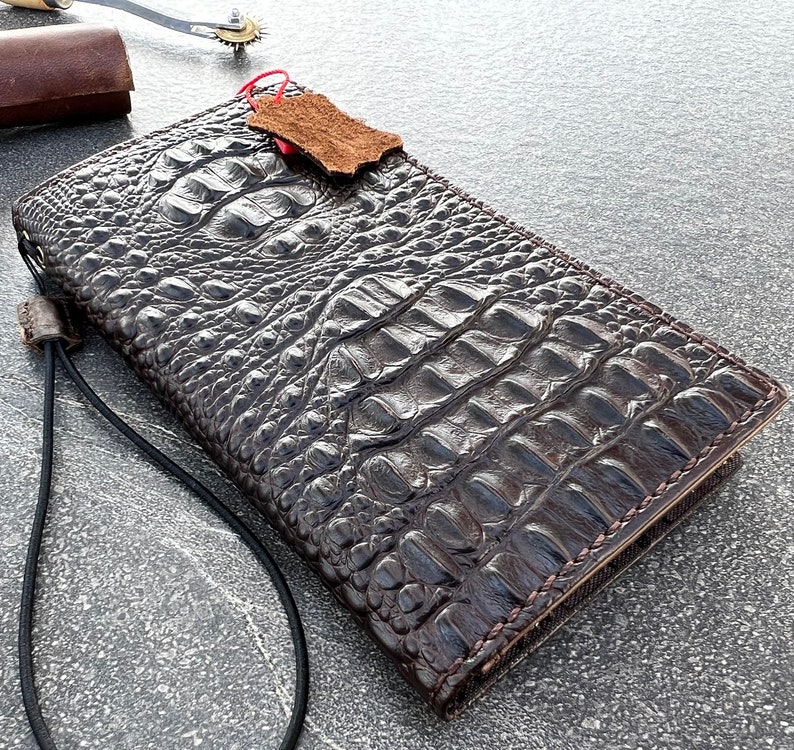 May include: A dark brown alligator skin wallet with a strap. The wallet has a textured surface and is made of high-quality leather.