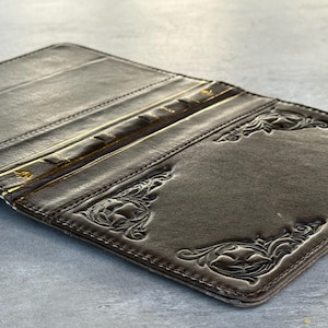 Peut inclure: Portefeuille en cuir marron ouvert avec des motifs floraux en relief aux coins. Le portefeuille comporte plusieurs fentes pour cartes et une bordure cousue. L'intérieur est d'une teinte plus claire que l'extérieur.