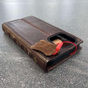 May include: A brown leather phone case designed to look like a book. The case has gold-colored accents and a red tag attached. The case is made of leather and has a flap that closes over the phone.