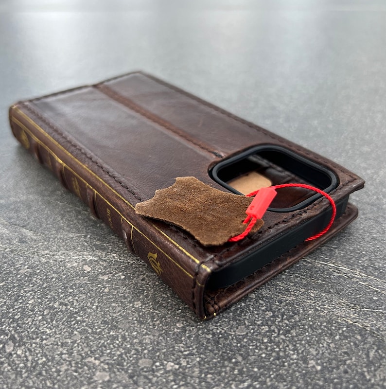 May include: A brown leather phone case designed to look like a book. The case has gold-colored accents and a red tag attached. The case is made of leather and has a flap that closes over the phone.