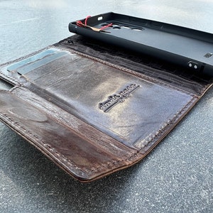 May include: A brown leather wallet with a phone case. The wallet is open, revealing card slots and a dark interior. The phone case is black and sits on top of the open wallet. The wallet has a distressed look.
