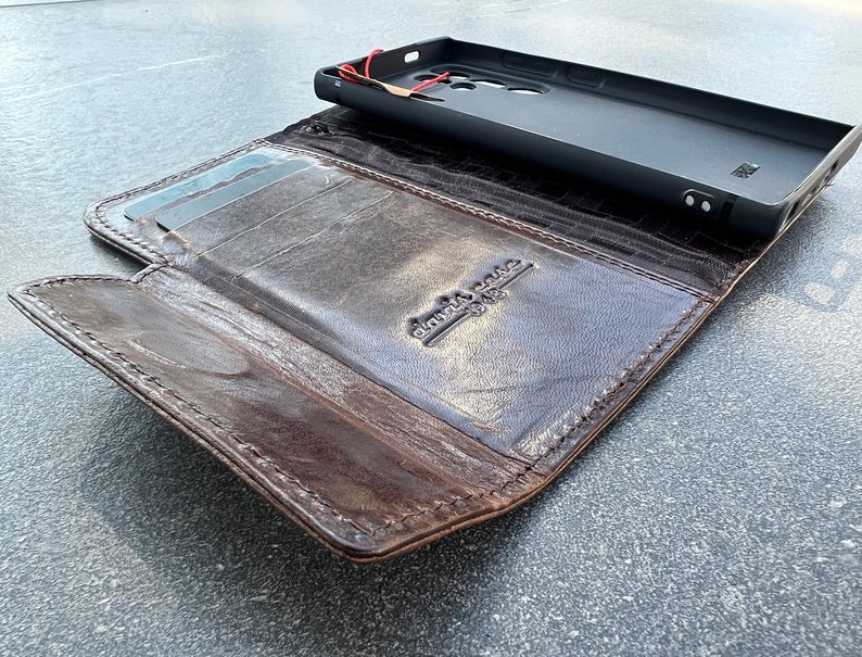 May include: A brown leather wallet with a phone case. The wallet is open, revealing card slots and a dark interior. The phone case is black and sits on top of the open wallet. The wallet has a distressed look.