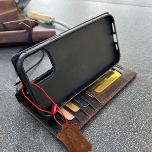 May include: A brown leather wallet case with a black phone case inside. The wallet has multiple card slots and a flap that closes with a snap. The phone case has a cutout for the camera lens. A red string is attached to the phone case.
