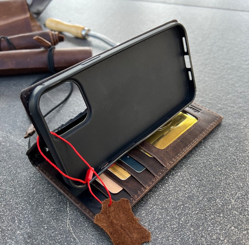 May include: A brown leather wallet case with a black phone case inside. The wallet has multiple card slots and a flap that closes with a snap. The phone case has a cutout for the camera lens. A red string is attached to the phone case.