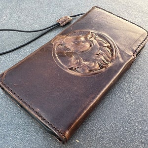 May include: A brown leather phone case with a horse head design embossed on the front. The case has a black strap attached to it.