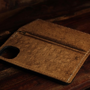 Puede incluir: Una funda de teléfono de cuero marrón con una superficie texturizada. La funda está abierta, revelando un interior negro y un recorte para la cámara. La funda está cosida con hilo blanco y descansa sobre una superficie de madera oscura.